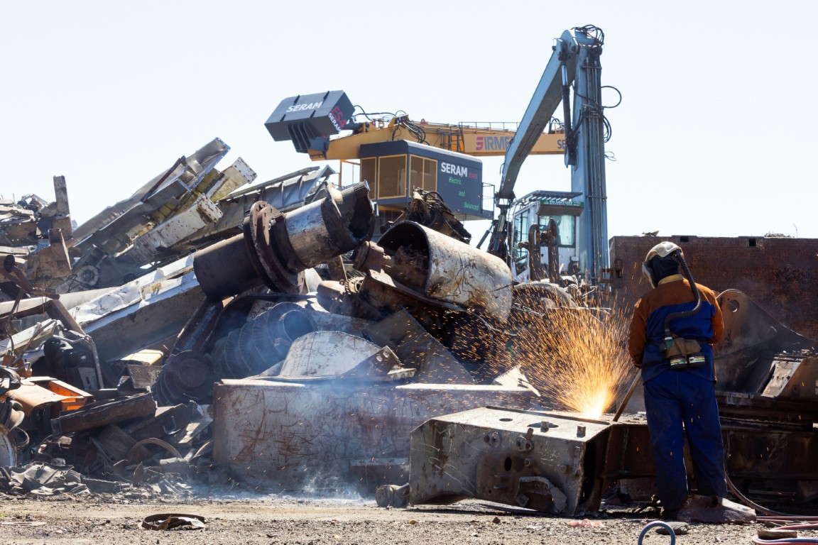 Chalumiste découpant une pièce métallique sur une plateforme de recyclage, avec grue et tas de ferrailles en arrière-plan